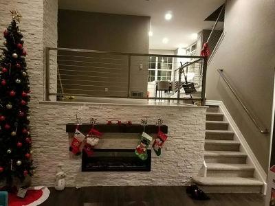 A festive modern living room features a white stone fireplace decorated with colorful Christmas stockings next to a tall, lit holiday tree and carpeted stairs leading to an upper level.