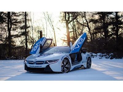 This image features a white BMW i8 sports car with its signature butterfly doors open, parked in a snow-covered field during a bright sunset. The vehicle is branded with blue and white "VIP" decals and a website URL, set against a background of bare win