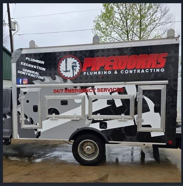 A professionally wrapped Pipeworks Plumbing & Contracting utility truck featuring the company's logo, services (Plumber, Excavation, General Contractor), "24/7 Emergency Services" banner in red, and open tool compartments, parked on a wet surface on an
